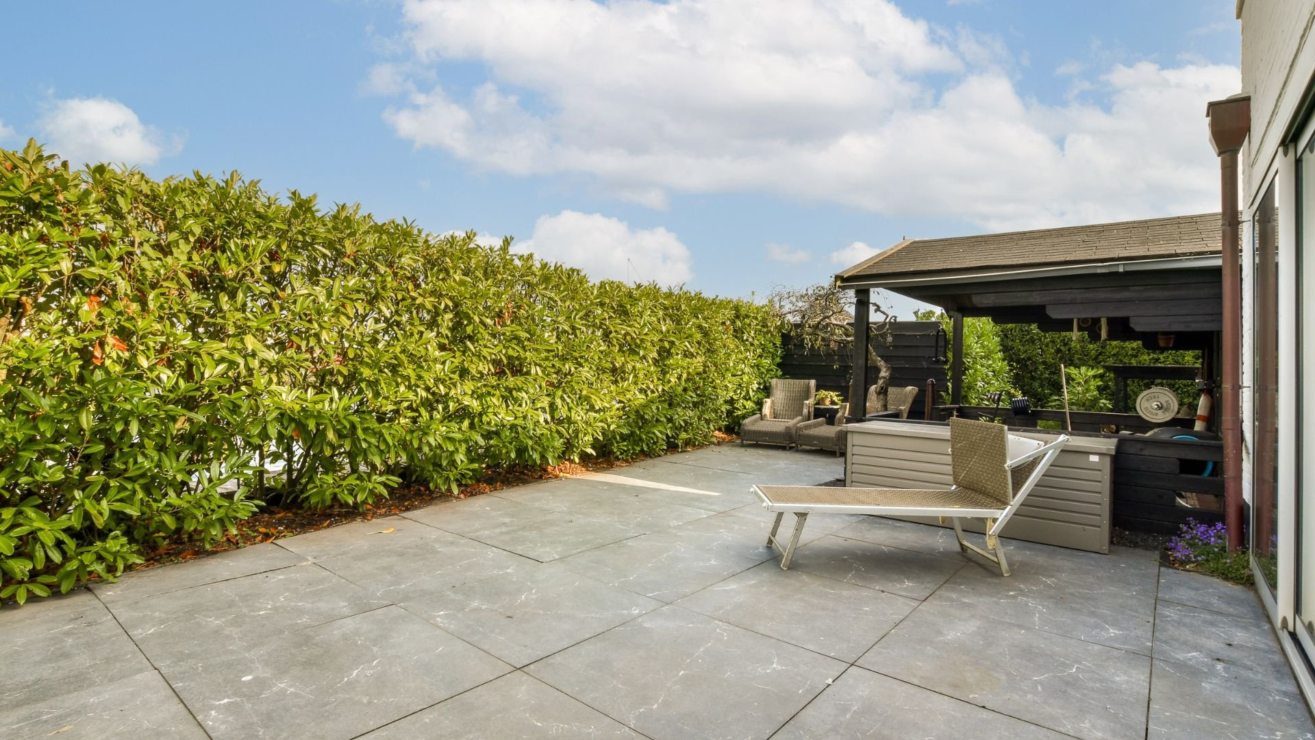 Outdoor patio with gray tiles, lounge chair, and green hedge backdrop